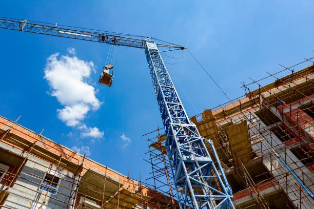 Shot from below of crane that puts down on ground wheelbarrow full of industrial waste, crumpled cellophane, in background is edifice under construction with scaffold and blue clear sky.